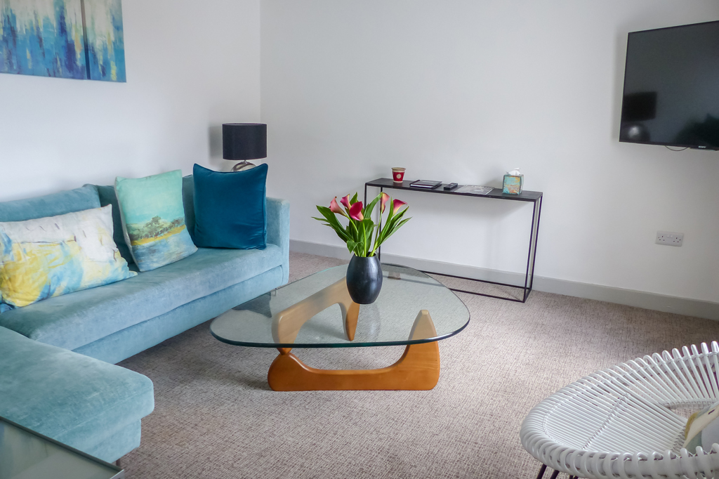 Living Area with Flatscreen Smart TV mounted to wall. L shaped Sofa pictured on the left side. Curved edged three sided glass topped coffee table with a plant pot in the centre of the shot.